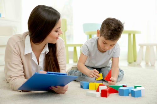 Woman watching a boy play with colorful building blocks on the floor.