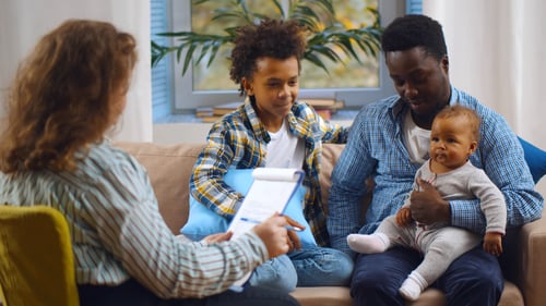 A man and two children sit on a sofa during a consultation with a person holding a clipboard.